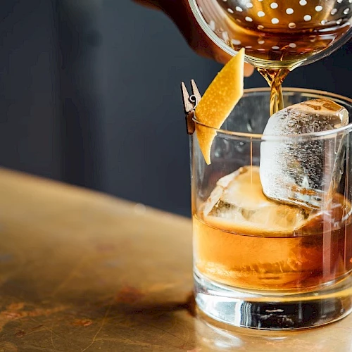 A glass with whiskey and a large ice cube, garnished with a slice of orange, as a bartender pours a drink into it, on a wooden bar.