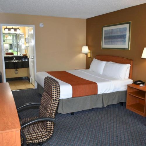 A hotel room with a bed, two nightstands, lamps, a desk and chair, an armchair, and an adjacent bathroom area, neat and functional.