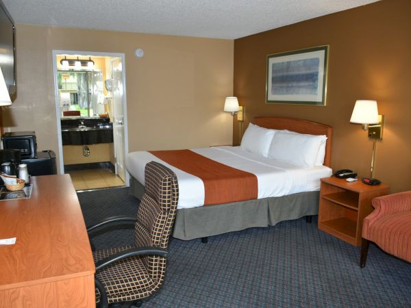 A hotel room with a queen bed, two nightstands, a desk and chair, a sofa, lamps, and an en-suite bathroom visible through an open door.