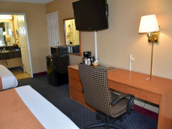 A hotel room with a desk, chair, TV on the wall, mini-fridge, coffee maker, mirror, bed, and attached bathroom area, lit by a lamp.