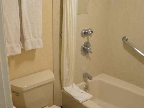 A small hotel bathroom with a toilet, bathtub, shower curtain, towel rack, and a grab bar, all in beige tones.
