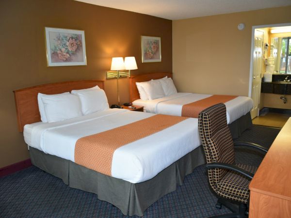 Two double beds with white linens and orange runners, a desk and chair, lamps, wall art, and a small sink area visible in a tidy hotel room.