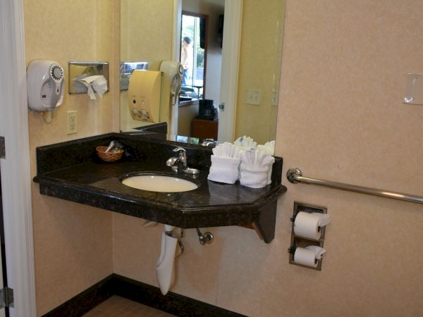 A hotel bathroom with a granite counter, sink, mirror, wall-mounted soaps, stacked towels, a toilet, and a grab bar by the wall.